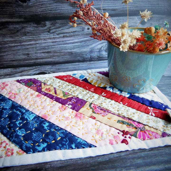 A handmade quilted fabric placemat with a colorful patchwork design of floral and patterned stripes, styled on a dark wooden table next to a light blue ceramic vase filled with dried flowers.